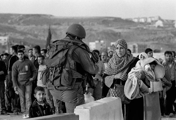 Territori occupati palestinesi. Palestina, Qalandia - Check Point dell'esercito israeliano (Israeli Defence Force) - Militare controlla donna con neonato in braccio e bambini - Gruppo di persone sullo sfondo