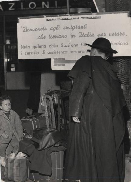 Milano - Stazione Centrale - Interno - Emigranti tornano per votare - Cartello di benvenuto - Bambino seduto su valigie - Prete