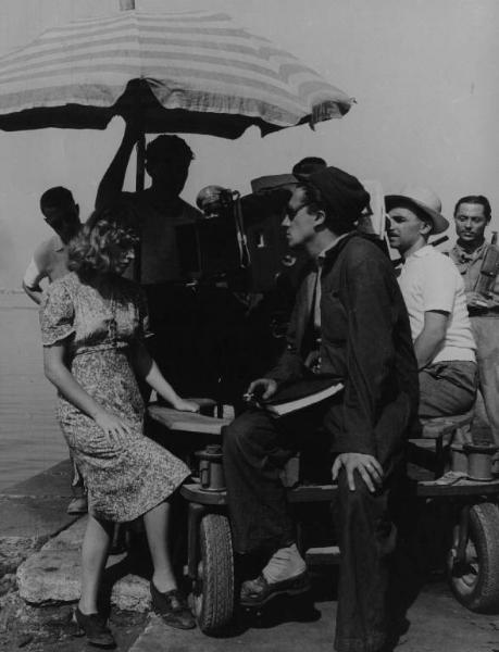 Set del film "Senza pietà" - Regia Alberto Lattuada - 1948 - L'attrice Carla Del Poggio sul set sotto l'obrellone e il regista Alberto Lattuada dietro la macchina da presa, attorno gli operatori della troupe