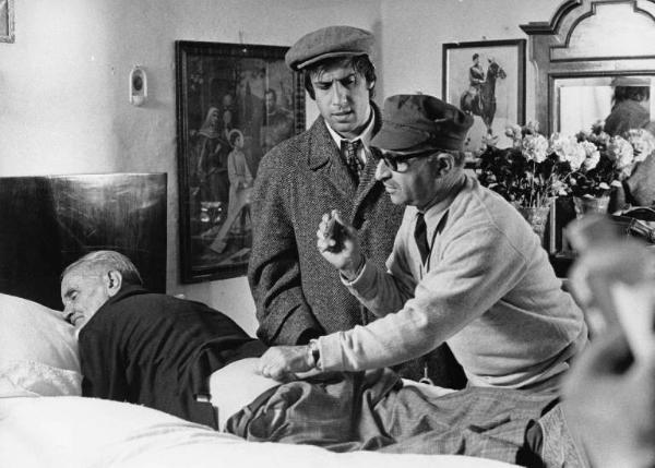 Set del film "Bianco, rosso e..." - Regia Alberto Lattuada - 1972 - Il regista Alberto Lattuada sul set mentre fa un'iniezione a un attore non identificato steso sul letto e l'attore Adriano Celentano e ,