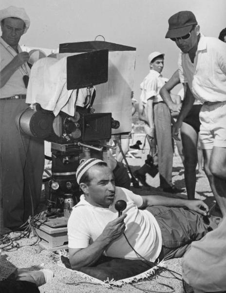 Set del film "La spiaggia" - Regia Alberto Lattuada - 1953 - Il regista Alberto Lattuada sdraiato sulla spiaggia parla al microfono davanti alla macchina da presa tra gli operatori della troupe