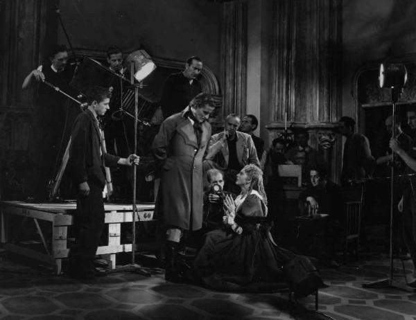 Set del film "Il Conte di Brechard" - Regia Mario Bonnard - 1938 - L'attore Amedeo Nazzari osserva l'attrice Luisa Ferida inginocchiata davanti a lui, mentre il regista Mario Bonnard e lo staff tecnico li dirigono