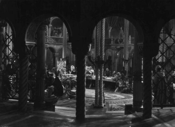 Set del film "La corona di ferro" - Regia Alessandro Blasetti - 1940 - L'attrice Elisa Cegani al tavolo con attori non identificati visti da una ambiente adiacente.