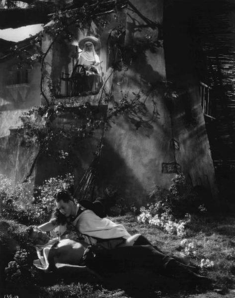 Set del film "La corona di ferro" - Regia Alessandro Blasetti - 1940 - L'attore Massimo Girotti è sdraiato a terra e l'attrice Luisa Ferida gli si avvicina, mentre l'attrice Rina Morelli dal balcone osserva la scena.