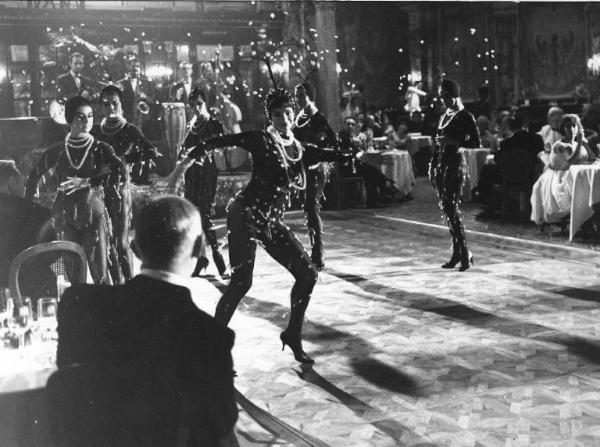Set del film "Crimen" - Regia Mario Camerini- 1960 - Un gruppo di attrici non identificate balla al centro di un salone dove attori non dientificati siedono a dei tavoli.