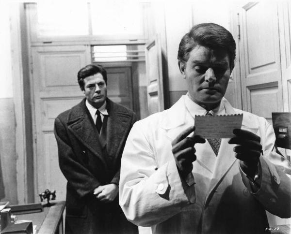 Set del film "Cronaca familiare" - Regia Valerio Zurlini 1962 - L'attore Marco Guglielmi nei panni del medico legge un foglietto in primo piano; alle sue spalle l'attore Marcello Mastroianni.