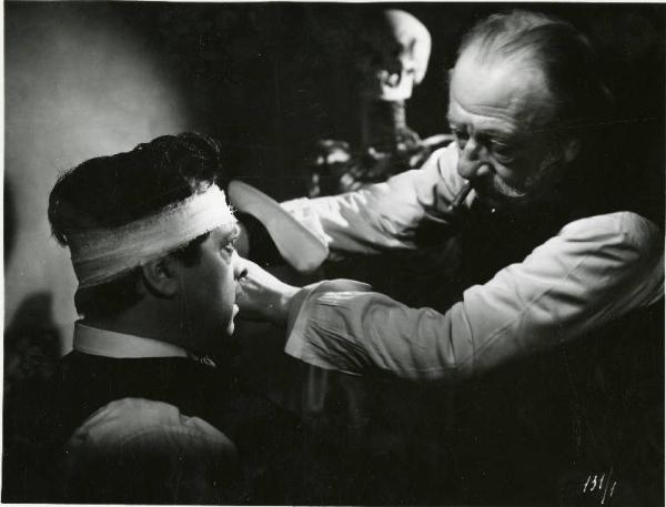 Scena del film "Il delitto di Giovanni Episcopo" - Regia Alberto Lattuada, 1947 - Un uomo anziano, con un sigaro in bocca, fascia la testa di Aldo Fabrizi con una benda. Sullo sfondo uno scheletro umano.