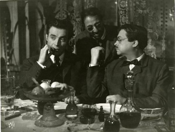 Scena del film "Il delitto di Giovanni Episcopo" - Regia Alberto Lattuada, 1947 - Roldano Lupi, seduto a una tavola imbandita, tiene un sigaro in bocca. Seduto accanto a lui, Aldo Fabrizi parla con un uomo, in piedi, alle sue spalle.