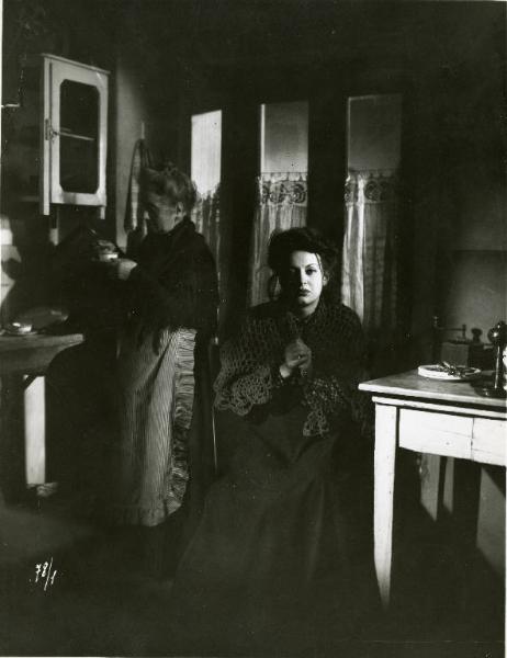 Scena del film "Il delitto di Giovanni Episcopo" - Regia Alberto Lattuada, 1947 - Yvonne Sanson seduta in cucina, spettinata con indosso uno scialle si stringe le mani davanti al petto. Sua madre, Ave Ninchi, prepara il caffè.