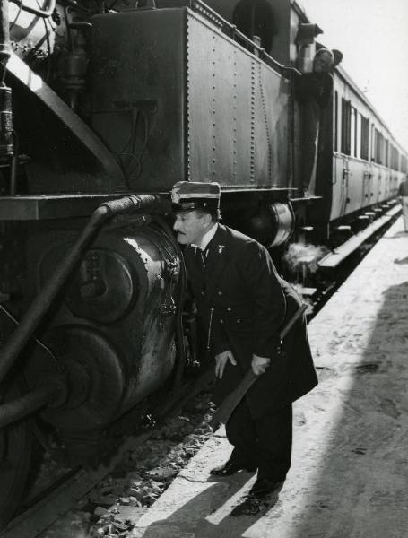 Scena del film "Destinazione Piovarolo" - Regia Domenico Paolella, 1955 - Totò, in piedi a tre quarti, accanto a un treno fermo, si china leggermente in avanti e guarda fisso verso di esso.