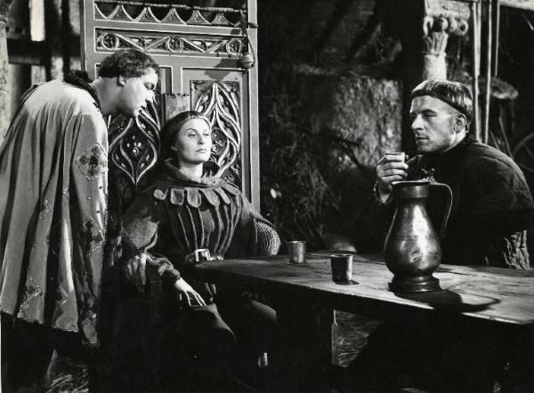 Scena del film "Destini di donne" - Regia Christian-Jaque, Jean Delannoy, Marcello Pagliero, 1954 - Michèle Morgan seduta su un trono alla testa di un tavolo. In piedi, a sinistra, Daniel Ivernel. A destra, seduto, un ragazzo.