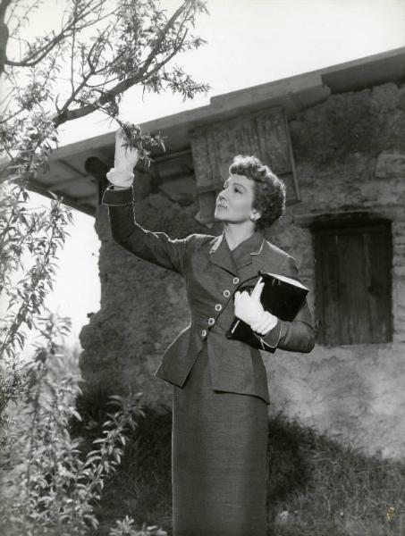 Scena del film "Destini di donne" - Regia Christian-Jaque, Jean Delannoy, Marcello Pagliero, 1954 - Piano americano di Claudette Colbert in piedi di fronte a un albero, tiene in mano un ramo e lo osserva. Nell'altra mano un quaderno.