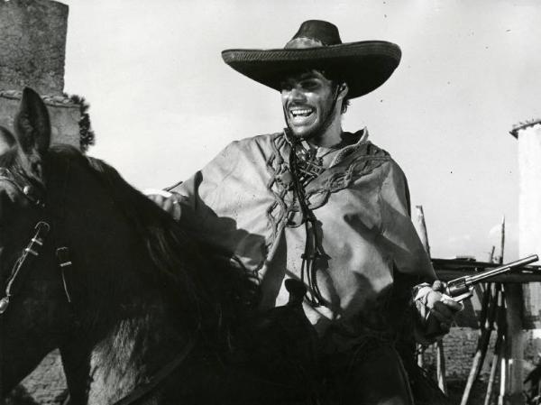 Scena del film "10.000 dollari per un massacro" - Regia Romolo Girolami, 1967 - Mezza figura di Claudio Camaso, con poncho e cappello, in sella a un cavallo mentre ride e impugna una pistola.