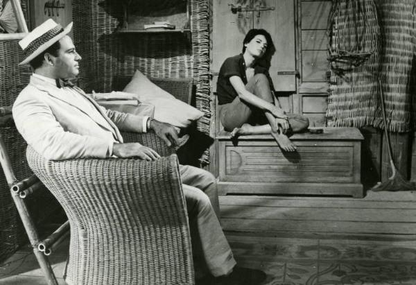 Scena del film "La diga sul Pacifico" - Regia René Clément, 1957 - In primo piano, di profilo, Nehemiah Persoff è seduto su una poltrona di vimini. Sullo sfondo, Silvana Mangano è seduta a gambe incrociate su una cassapanca.