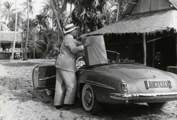 Scena del film "La diga sul Pacifico" - Regia René Clément, 1957 - Nehemiah Persoff, regge con le mani un pacco appoggiato a una decappottabile. L'attore ha la lingua fuori dalla bocca.