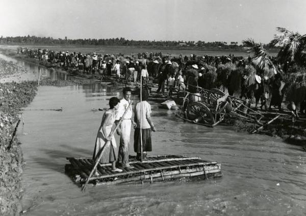 Scena del film "La diga sul Pacifico" - Regia René Clément, 1957 - In mezzo a un corso di acqua, in piedi su una zattera Silvana Mangano, Richard Conte e Jo Van Fleet. La Mangano rema. Sullo sfondo, una lunga fila di persone.