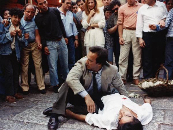 Scena del film "Dimenticare Palermo" - Regia Francesco Rosi, 1990 - James Belushi è accovacciato accanto al cadavere di Marco Leonardi. Sullo sfondo la folla assiste alla scena.