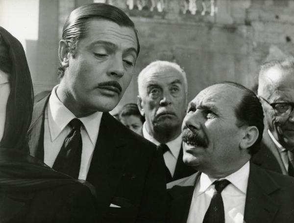 Scena del film "Divorzio all'italiana" - Regia Pietro Germi, 1961 - Marcello Mastroianni in primo piano ascolta un uomo accanto a lui. Dietro di loro, Odoardo Spadaro e altre persone in fila per il corteo funebre.