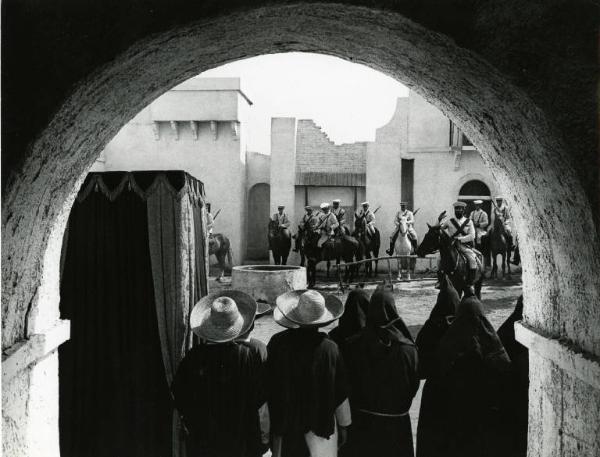 Scena del film "Un dollaro tra i denti" - Regia Luigi Vanzi, 1966 - Sotto un arco, un gruppo di uomini, tra cui dei monaci, sono in piedi, di spalle. Sullo sfondo, dei soldati a cavallo.