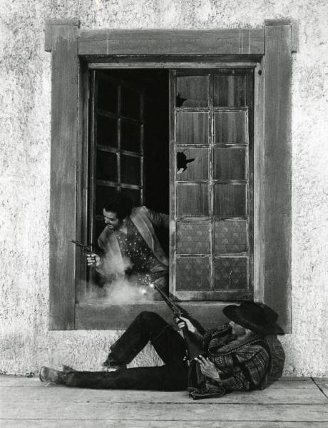 Scena del film "Un dollaro tra i denti" - Regia Luigi Vanzi, 1966 - Totale. Un uomo, sdraiato sotto una finestra, spara a un altro che si affaccia alla stessa.