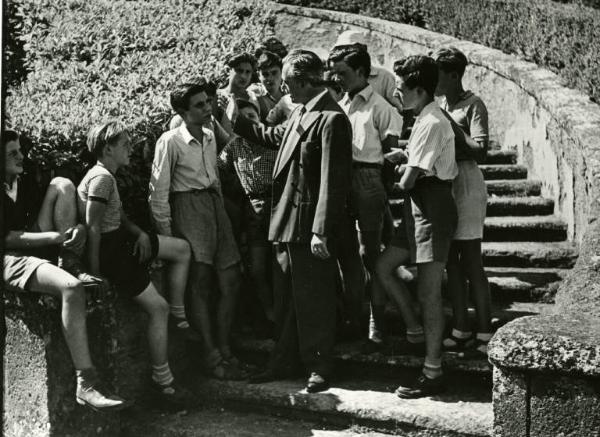 Scena del film "Domani è troppo tardi" - Regia Léonide Moguy, 1950 - Totale. Esterno. Ai piedi di una scalinata Vittorio De Sica, di profilo, parla a un gruppo di ragazzi attorno a lui.