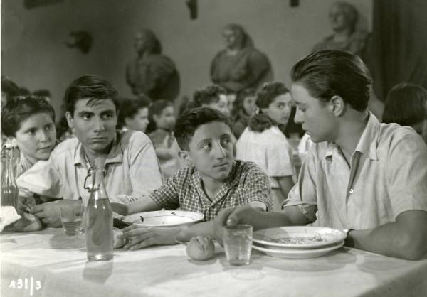 Scena del film "Domani è troppo tardi" - Regia Léonide Moguy, 1950 - Carlo Delle Piane è seduto a tavola con altri ragazzi. Alla sua sinistra, Gino Leurini. Alle sue spalle siede Anna Maria Pierangeli con il viso e lo sguardo rivolto verso di lui.