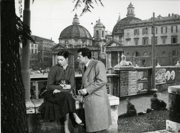 Scena del film "Donatella" - Regia Mario Monicelli, 1956 - Totale. Elsa Martinelli, seduta su un muretto, tiene in braccio un cagnolino. Al suo fianco, Gabriele Ferzetti, in piedi, le parla.