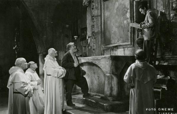 Scena del film "Don Buonaparte" - Regia Flavio Calzavara, 1941 - Totale. All'interno di una chiesa accanto a un altare quattro monaci e un uomo guardano verso l'alto, dove un altro uomo, in piedi su una scala a pioli, legge un documento.