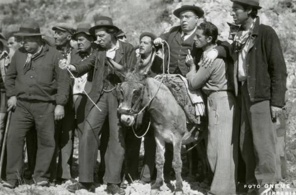 Scena del film "Don Buonaparte" - Regia Flavio Calzavara, 1941 - Totale. Un uomo, con occhiali e cappello in sella a un asino, si appoggia timoroso a un altro. Accanto a loro, a sinistra, Aldo Silvani e altri uomini.