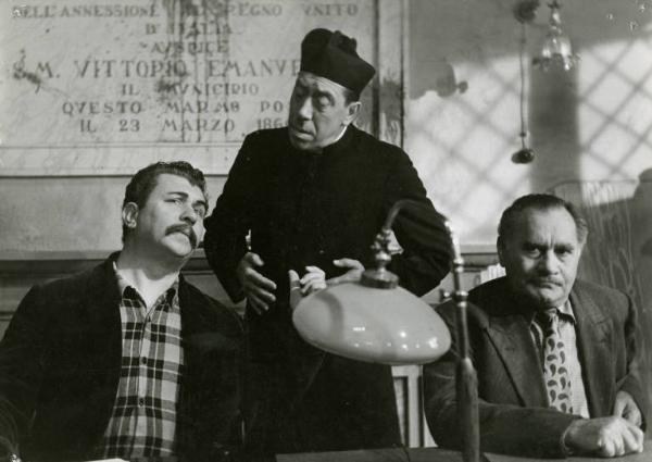 Scena del film "Don Camillo" - Regia Julien Duvivier, 1952 - Campo medio. Gino Cervi seduto al tavolo del Consiglio Comunale. Dietro di lui, in piedi, Fernandel, lo guarda. A destra, un attore non identificato.