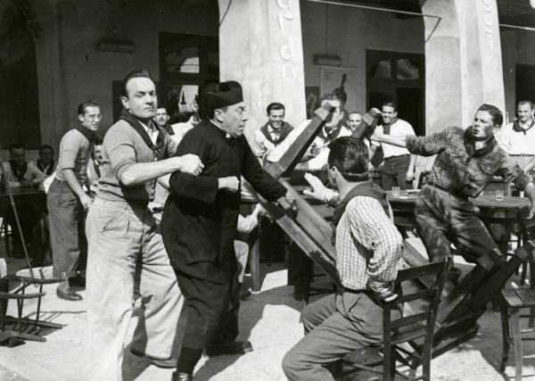 Scena del film "Don Camillo" - Regia Julien Duvivier, 1952 - Totale. Fernandel rovescia un tavolo mentre un uomo alle sue spalle cerca di trattenerlo. Sullo sfondo un gruppo di ragazzi assiste divertito alla scena.