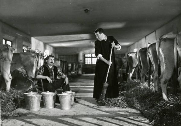 Scena del film "Don Camillo" - Regia Julien Duvivier, 1952 - Totale. All'interno di una stalla Fernandel spazza e Gino Cervi siede su uno sgabello da mungitore. I due si guardano.