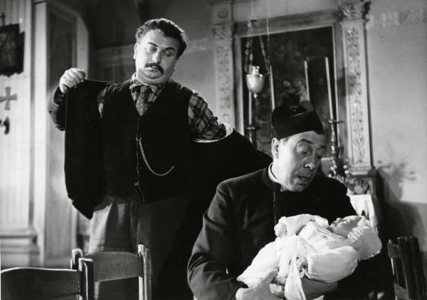 Scena del film "Don Camillo" - Regia Julien Duvivier, 1952 - Mezza figura di Fernandel seduto mentre culla un neonato. Dietro di lui in piedi, Gino Cervi, si infila la giacca.