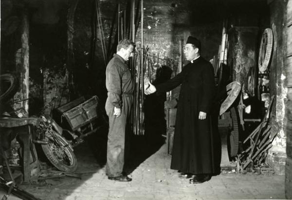 Scena del film "Don Camillo" - Regia Julien Duvivier, 1952 - Figura intera di Fernandel mentre tende la mano a Gino Cervi, in tuta da meccanico, che la guarda.