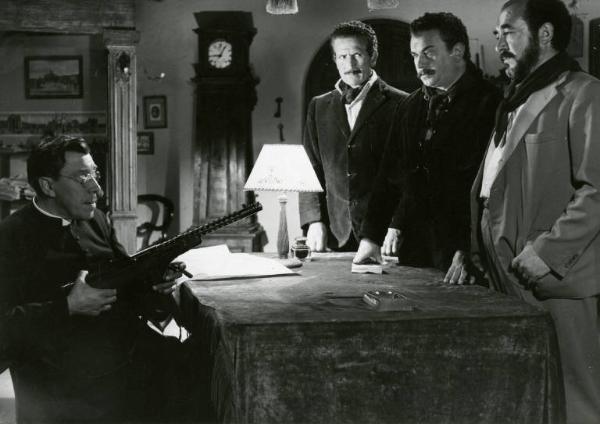 Scena del film "Don Camillo" - Regia Julien Duvivier, 1952 - Fernandel, seduto dietro una scrivania, minaccioso punta un mitra. Di fronte a lui Saro Urzì, Gino Cervi e un attore non identificato lo guardano.