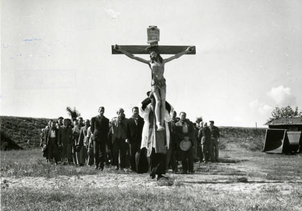 Scena del film "Don Camillo" - Regia Julien Duvivier, 1952 - Esterno. Fernandel porta un crocifisso tenendolo in verticale di fronte a sé. Dietro di lui, una piccola folla, in processione.