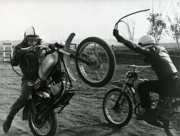 Scena del film "Don Camillo e i giovani d'oggi" - Regia Mario Camerini, 1972 - Due motociclisti, in sella alle loro motociclette, si fronteggiano. Uno impenna la sua moto mentre l'altro gli va incontro, agitando in aria una cinghia con catena.