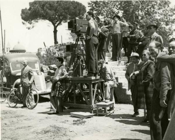 Sul set del film "Don Giovanni" - Regia Dino Falconi, 1942 - Totale. Due operatori dietro alla macchina da presa posta su un piccolo trabattello quadrato. Alle loro spalle un gruppo di persone in piedi assiste alla scena.