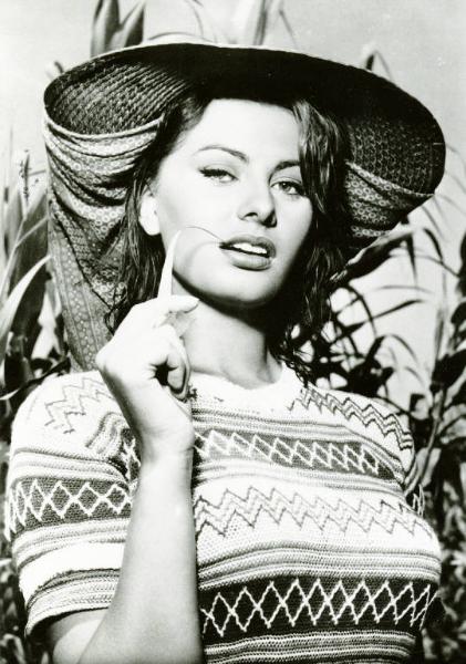 Scena del film "La donna del fiume" - Regia Mario Soldati, 1955 - Mezza figura frontale di Sophia Loren. L'attrice indossa un cappello e con la mano destra porta un filo d'erba allo spigolo della bocca socchiusa.