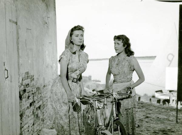 Scena del film "La donna del fiume" - Regia Mario Soldati, 1955 - Piano americano di Sophia Loren, a sinistra e Lise Bourdin, a destra. Le attrici, tengono ognuna la propria bicicletta, per il manubrio. Sullo sfondo, il fiume.