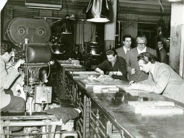 Sul set del film "La donna del giorno" - Regia Francesco Maselli, 1957 - Mario Carotenuto e Antonio Cifariello, dietro di lui, passano dietro un banco di lavoro dove due uomini stanno lavorando. Sulla sinistra, un operatore, li riprende.