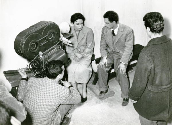 Sul set del film "La donna del giorno" - Regia Francesco Maselli, 1957 - Francesco Maselli, seduto su una panca, tiene le mani sulle ginocchia e guarda una donna seduta su una sedia, accanto a lui. Un operatore accovacciato, li riprende.