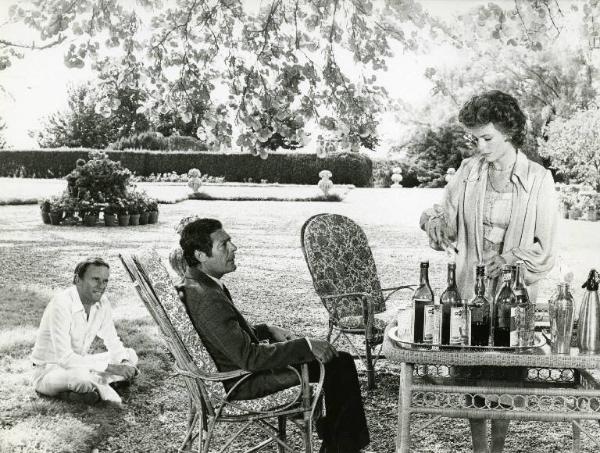 Scena del film "La donna della domenica" - Regia Luigi Comencini, 1975 - In un giardino Marcello Mastroianni, seduto su una sedia, di profilo, guarda Jacqueline Bisset, in piedi, che prepara dei cocktails. Jean-Louis Trintignant è seduto per terra.