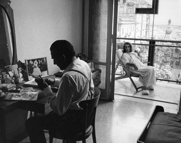 Scena del film "La donna scimmia" - Regia Marco Ferreri, 1964 - In primo piano, Ugo Tognazzi, seduto a una scrivania, taglia delle foto. Sullo sfondo, Annie Girardot, sul balcone, seduta su una sdraio, lo osserva.