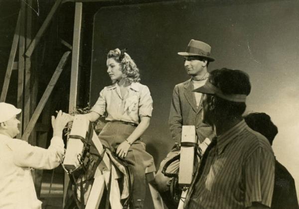 Sul set del film "Dora Nelson" - Regia Mario Soldati, 1939 - Assia Noris e un attore non identificato siedono sopra finti cavalli di legno. Attorno a loro, uomini della troupe.