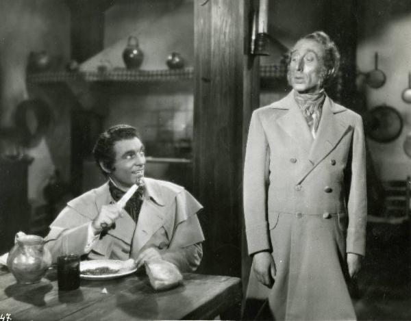 Scena del film "Il dottor Antonio" - Regia Enrico Guazzoni, 1937 - Mino Doro, seduto a tavola, porta alla bocca un coltello con infilzato del cibo e sorride a un attore non identificato in piedi accanto a lui.