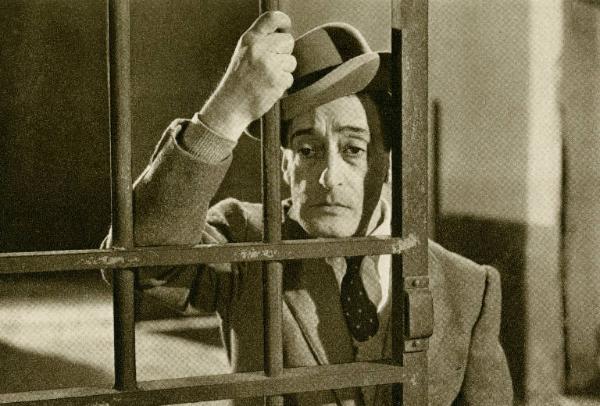 Scena del film "Dov'è la libertà?" - Regia Roberto Rossellini, 1954 - Mezza figura di Totò che tiene il mento appoggiato alle sbarre di una cella aperta, alle quali si aggrappa con una mano.