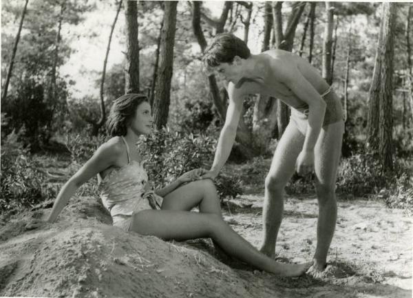 Scena del film "Domenica d'agosto" - Regia Luciano Emmer, 1950 - In una pineta, Anna Baldini è seduta su un mucchio di sabbia e Franco Interlenghi, in piedi di fronte a lei, le posa una mano sul ginocchio. Entrambi sono in costume da bagno.