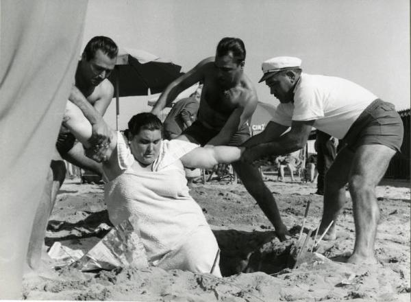 Scena del film "Una domenica d'estate" - Regia Giulio Petroni, 1962 - Totale. In spiaggia, tre uomini aiutano una donna obesa a uscire da una buca.