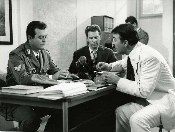 Scena del film "Una domenica d'estate" - Regia Giulio Petroni, 1962 - A sinistra, un poliziotto, dietro una scrivania, interroga Franco Fabrizi, seduto di fronte a lui. Seduto tra loro, un attore non identificato alza la cornetta del telefono.